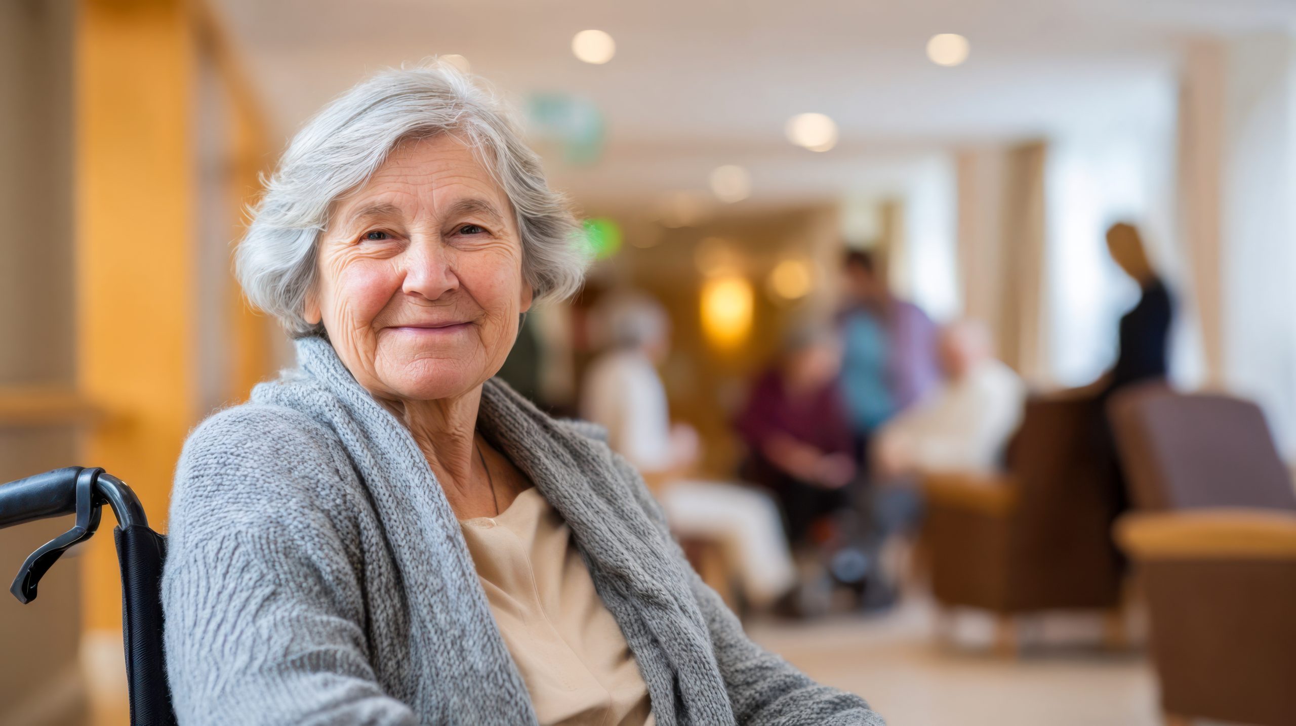 Older woman smiles while sitting in a wheelchair in a care facility during daytime activities with other residents in the background lady smiling